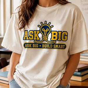 Può includere: Una t-shirt a maniche corte color bianco sporco con una stampa grafica raffigurante una lampadina blu e gialla. Il testo ASK BIG è scritto in grandi lettere gialle con bordi blu, e sotto, un banner blu riporta ASK BIG • BUILD SMART.
