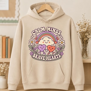May include: A beige hooded sweatshirt featuring a graphic print of a smiling cloud, a rainbow, and two anthropomorphic hearts holding hands. The text "CALM MINDS, BRAVE HEARTS" is printed in a curved font around the central illustration.