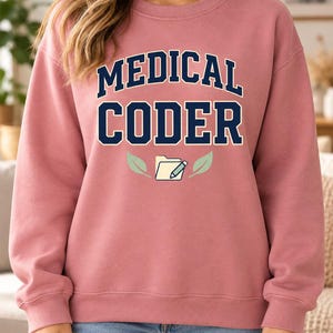 Peut inclure: Un sweat-shirt à col rond rose poudré avec le texte bleu foncé MEDICAL CODER dans une police universitaire. Sous le texte se trouve un graphique d'un dossier jaune clair avec un crayon bleu, entouré de deux feuilles vert clair.