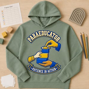 May include: A sage green hooded sweatshirt featuring a graphic print of hands stacking blue and yellow blocks with a pencil. The text reads "PARAEDUCATOR" in an arch above and "PATIENCE IN ACTION" on a banner below the illustration.