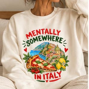 Peut inclure: Un sweat-shirt à col rond couleur crème arborant une impression graphique colorée avec le texte "MENTALLY SOMEWHERE IN ITALY". Le motif comprend une part de pizza, des citrons jaunes et un village côtier italien avec des cœurs rouges.