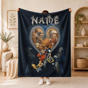 Peut inclure: Une couverture en polaire bleu foncé ornée d'un grand motif en forme de cœur représentant des personnages de Kingdom Hearts, dont Sora, Donald et Riku. Le mot NAME est imprimé dans une police gothique stylisée au-dessus de l'emblème.