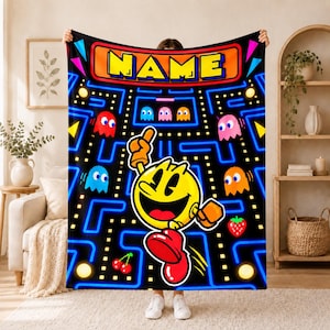 Può includere: Una coperta in pile nera con un vivace design a labirinto Pac-Man, pareti blu neon, fantasmi colorati, icone di frutta e un grande personaggio Pac-Man giallo al centro. La parte superiore presenta un banner rettangolare arancione con il testo NAME.