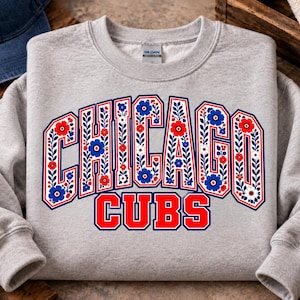 Puede incluir: Una sudadera gris claro de cuello redondo con el texto CHICAGO CUBS en una fuente de letras de bloque. La palabra CHICAGO está rellena con un patrón decorativo de flores y hojas rojas, azules y blancas, mientras que CUBS es de color rojo sólido.