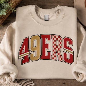 Op de afbeelding: Een zandkleurige sweater met ronde hals en de tekst 49ERS in een vet, gebogen lettertype. De cijfers en letters zijn gekleurd in rood en goud, waarbij de letter R een rood-wit geblokt patroon heeft met een gouden omlijning.