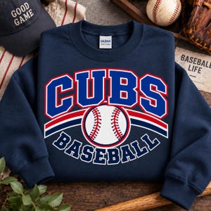Puede incluir: Una sudadera azul marino de cuello redondo con un estampado gráfico que muestra el texto CUBS en letras grandes azules con bordes blancos y rojos. Debajo del texto hay una ilustración de una pelota de béisbol blanca con costuras rojas y la palabra BASEBALL.
