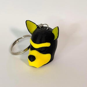 Peut inclure: Un porte-clés imprimé en 3D représentant une tête d'animal stylisée avec une base noire et des accents jaune vif sur les oreilles, le front et le museau. Le design comprend un nez noir et un anneau fendu en métal argenté fixé au sommet.