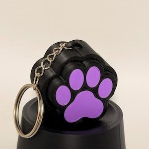 May include: A black 3D printed keychain shaped like a paw print with vibrant purple pads. The object features a textured layered surface and is attached to a silver metal split ring by a short chain, creating a functional accessory for keys.