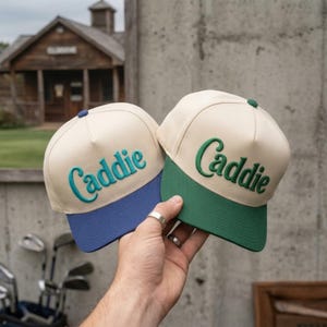 May include: Two baseball caps with cream-colored crowns and structured panels. One cap features a blue brim and teal embroidered text reading Caddie, while the other has a green brim and dark green embroidered text reading Caddie.
