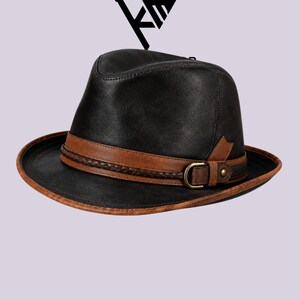 May include: A black fedora hat featuring a brown leather band with a braided texture, a side buckle with a metallic D-ring, and a matching brown trim along the edge of the brim. The hat has a structured crown and a wide, slightly curved brim.