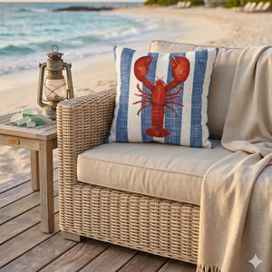 May include: A square throw pillow featuring a vibrant red lobster illustration centered on a background of vertical blue and white stripes. The pillow is placed on a light beige outdoor sofa cushion, accompanied by a matching beige throw blanket.