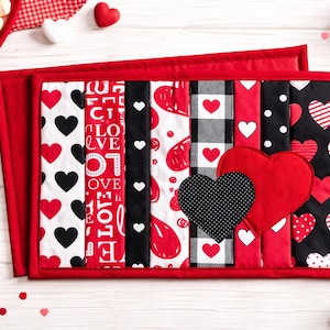 May include: A rectangular quilted placemat featuring vertical fabric strips in red, black, and white with various heart patterns, text reading LOVE, buffalo plaid, and polka dots, accented by two large fabric heart appliqués in red and black.