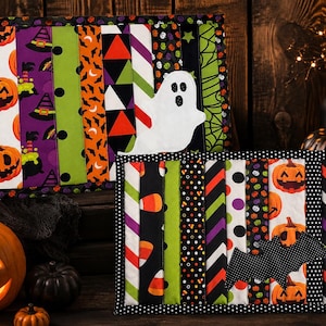 May include: Two rectangular quilted fabric mats featuring vertical strips of Halloween-themed patterns including pumpkins, candy corn, and spider webs. One mat displays a white ghost appliqué, while the other features a black bat appliqué.