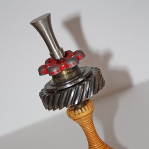 May include: A wooden base with a metal gear and red valve on top. The gear is attached to a metal pipe with a silver cap.