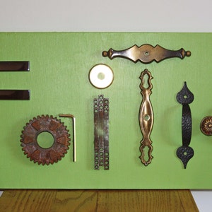 May include: A green wooden board with various metal hardware pieces arranged to spell the word "FAITH". The hardware includes door knobs, pulls, hinges, and a gear.