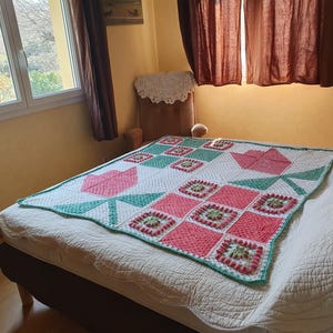 Peut inclure: Une couverture en crochet faite à la main présentant un motif patchwork de tulipes roses et blanches, des feuilles vertes et de petits carrés granny avec du fil rose, vert et blanc. La couverture a une bordure blanche et est posée sur un lit.