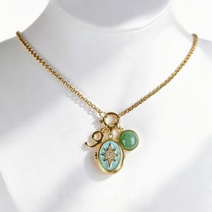 May include: A gold-tone chain necklace featuring a central oval pendant with a light blue enamel background and a central star motif set with small clear stones. The necklace also includes a small round green stone charm and a gold-tone swirl charm.