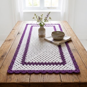 May include: A rectangular crochet table runner featuring a white center with a repeating granny square pattern, bordered by a thick purple frame and a scalloped purple edge. The runner is placed on a wooden table with a ceramic vase and bowl.