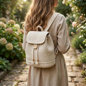 May include: A cream-colored crochet backpack featuring a structured flap, a single wooden button closure, and two drawstring cords with small wooden bead accents at the ends. The backpack has wide, matching crochet shoulder straps.
