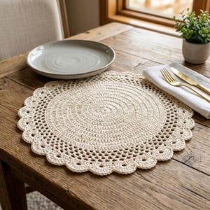 May include: A round, cream-colored crochet placemat featuring a dense, circular center pattern that transitions into an openwork mesh design, finished with a scalloped edge. The placemat is displayed alongside a plain ceramic plate and silverware.
