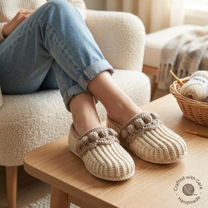 May include: A pair of cream-coloured crocheted slippers featuring a ribbed texture and a decorative brown crocheted trim with small bobble details along the top edge. The slippers are worn on feet clad in blue denim jeans, showcasing a slip-on design.