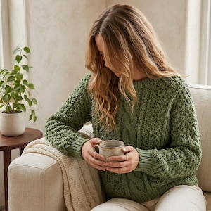 Può includere: Un maglione a maniche lunghe color verde salvia caratterizzato da un motivo a maglia testurizzato con dettagli a treccia verticali sul davanti. Il maglione presenta un girocollo e polsini a coste sui polsi e sull'orlo della struttura.