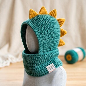May include: A teal crochet balaclava hood featuring a row of yellow triangular spikes along the top and back. The hood has a wide face opening and a ribbed neck section. A small white rectangular tag is attached to the lower front with text.