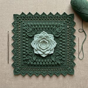 Può includere: Un quadrato all'uncinetto in verde bosco scuro con un motivo floreale tridimensionale centrale in verde menta chiaro, circondato da motivi a foglia testurizzati e un bordo smerlato decorativo, accompagnato da un uncinetto in metallo e filato.