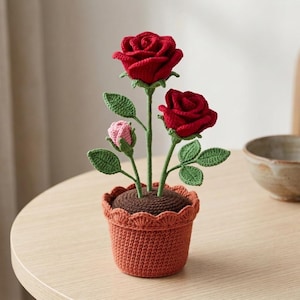 May include: A crochet potted plant featuring two large deep red roses and one small light pink rosebud on green stems with textured leaves. The plant is housed in a terracotta-coloured crocheted pot with a scalloped top edge and brown soil.