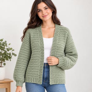 Può includere: Un cardigan aperto color verde salvia caratterizzato da un motivo a uncinetto testurizzato, maniche lunghe con polsini a coste e un orlo a coste. Il capo è indossato sopra un top bianco semplice e jeans blu, mostrando un punto di cucitura uniforme.