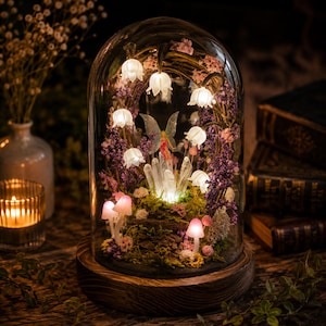 May include: A glass cloche dome on a circular wooden base containing a miniature scene with a small fairy figurine, clear quartz crystals, glowing white lily-shaped lights, and pink mushroom-shaped lights nestled in green moss and purple flowers.