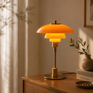 May include: A table lamp featuring a tiered, bright orange shade with three concentric layers. The lamp has a metallic gold-toned stem and a circular base. The light is illuminated, casting a warm glow from beneath the stacked orange shades.