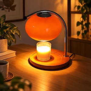 Könnte beinhalten: Eine Kerzenwärmerlampe mit einem leuchtend orangefarbenen Glaskuppelschirm, der von einem geschwungenen, verchromten Metallarm gehalten wird. Die Lampe steht auf einem runden Holzsockel, auf dem eine brennende weiße Kerze in einem Glas steht.