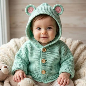 May include: A light mint green crocheted hooded cardigan featuring two small rounded ears on top with pink centers. The garment has long sleeves and three light brown wooden buttons down the front. The hem displays a textured cross-stitch pattern.