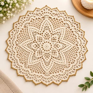 May include: A round, off-white crochet doily featuring an intricate, star-shaped floral pattern in the centre. The circular piece is finished with a delicate, scalloped edge highlighted by a thin, metallic gold thread trim along the entire border.