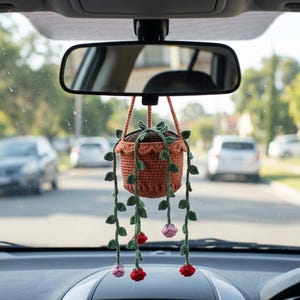 May include: A crocheted hanging plant ornament featuring a terracotta-colored pot with a scalloped rim. Long green vines with small leaves drape downwards, accented by several small, round, red and light pink crocheted flower blossoms.