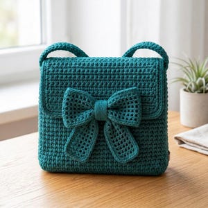 May include: A teal crochet handbag featuring a structured rectangular body and a prominent, matching teal crocheted bow attached to the front flap. The bag includes two short, rounded top handles and is crafted with a dense, uniform stitch pattern.