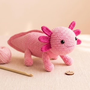Peut inclure: Un axolotl en peluche crocheté rose clair avec des branchies externes rose foncé, des yeux en plastique noir et une longue queue. La créature est réalisée avec des points de fil visibles et est placée à côté d'un crochet en bois et d'une étiquette.