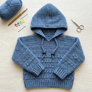 May include: A blue crocheted hooded sweatshirt featuring a textured bobble stitch pattern on the body and sleeves, long sleeves with ribbed cuffs, a hood with a drawstring, and two black toggles at the ends of the drawstring cords.