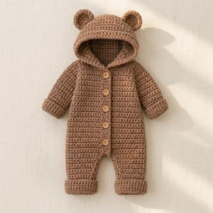 May include: A light brown crocheted one-piece garment featuring a hood with two small rounded bear ears on top. The garment has long sleeves, cuffed legs, and a front vertical opening secured by five round wooden buttons down the center.