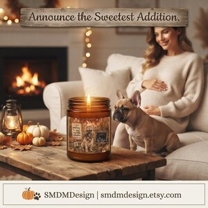 May include: A brown glass candle jar featuring a label with a French bulldog illustration, autumn leaves, and the text "I am waiting to meet my little pumpkin." The candle has a lit wick and sits on a wooden table in a cozy indoor setting.