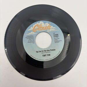 May include: A black 45 RPM vinyl record with a light blue center label featuring the Clouds logo, the song title Tip Toe To The Gas Pumps, and the artist name Tiny Tim. The label includes production and distribution text details.