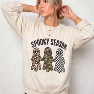 May include: A beige crewneck sweatshirt featuring the black text SPOOKY SEASON above three ghost illustrations. The first ghost has a brown and black leopard print, the second has a green and brown camouflage pattern, and the third has a black and white checkerboard pattern.