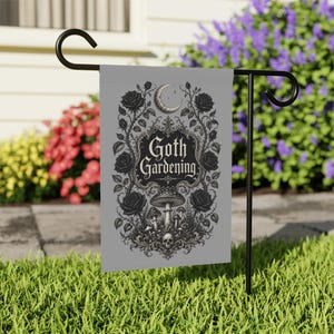 Könnte beinhalten: Eine rechteckige graue Gartenflagge an einem schwarzen Metallständer. Die Flagge zeigt eine schwarze Tuschezeichnung von Rosen, Pilzen, einem Totenkopf und einer Mondsichel, mit dem Text Goth Gardening in einem dekorativen Rahmen.