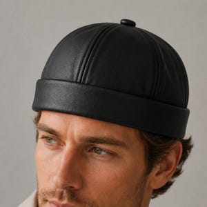 May include: A black leather docker cap featuring a rounded crown with vertical stitched paneling and a wide, folded brim. The hat has a smooth, matte texture and a small circular button detail at the very top centre of the dome-shaped crown.