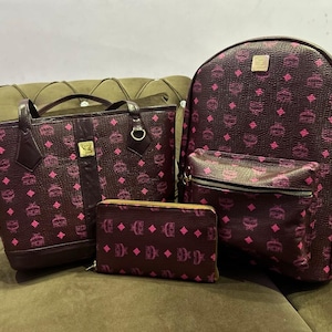 Puede incluir: Un bolso tote de color burdeos oscuro, una mochila a juego con bolsillo frontal con cremallera y una cartera rectangular. Cada artículo presenta un patrón de logotipo MCM repetido en rosa brillante, herrajes dorados y textura.