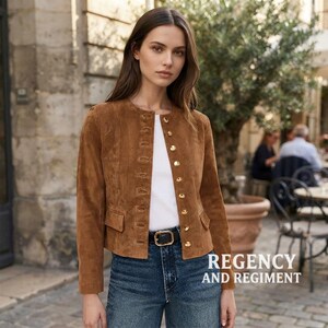 May include: A brown suede jacket featuring a round neckline, long sleeves, and a front button closure with gold-toned buttons. The jacket includes two small flap pockets at the waist and is worn over a plain white top and blue denim jeans with a black belt.