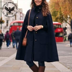 May include: A dark navy blue wool-blend cape coat featuring a high collar, a front button closure with metallic buttons, and two large patch pockets. The garment is worn over a black long-sleeved top and paired with a brown leather shoulder bag.