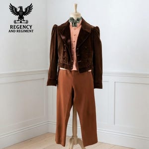 Puede incluir: Una chaqueta de terciopelo marrón oscuro de manga larga con cierre de doble botonadura, sobre una camisa abotonada de color melocotón claro con pajarita verde salvia y pantalones marrones a juego. El logo Regency and Regiment es visible.