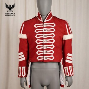 May include: A red military-style jacket featuring white braided cord detailing across the front, shoulders, and cuffs. The garment includes a high collar, silver-toned buttons, and white fur-like trim accents on the upper sleeves and shoulders.